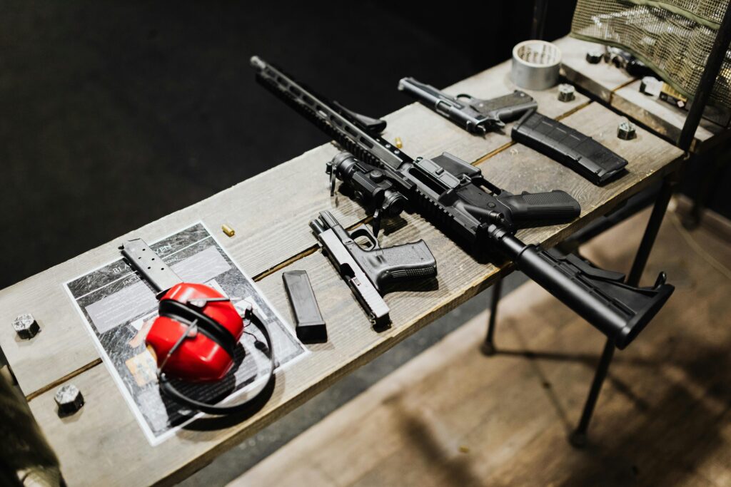 A variety of guns and accessories displayed on a wooden table indoors, featuring ear protection.